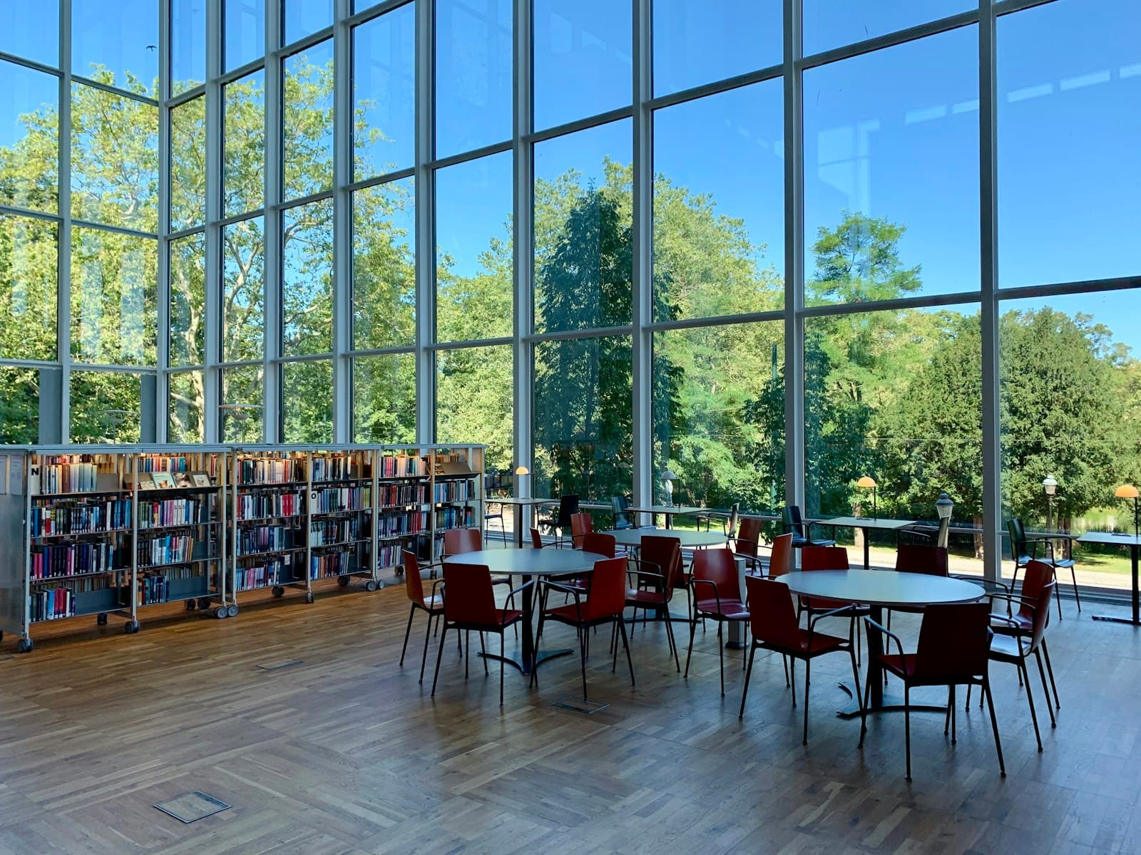 A bright library with tall windows, bookshelves and red chairs at round tables